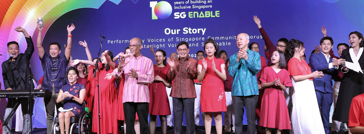 Group on stage in red dresses and patterned shirts clapping, with rainbow and "SG Enable" in background.